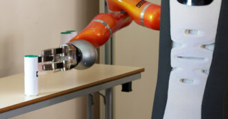 Orange robot arm with a silver three-fingered gripper on a Care-O-bot service robot, reaching for a small white canister with a green cap on a table
