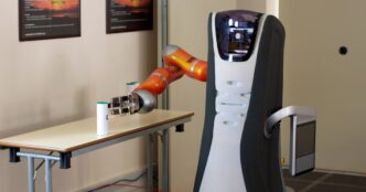 Orange robot arm with a silver three-fingered gripper on a Care-O-bot service robot, reaching for a small white canister with a green cap on a table