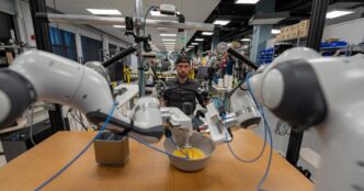 Toyota Research Institute bimanual robot with two white robot arms whisking eggs in a bowl on a wooden table while a researcher behind it holds teleoperation controllers in a robotics lab