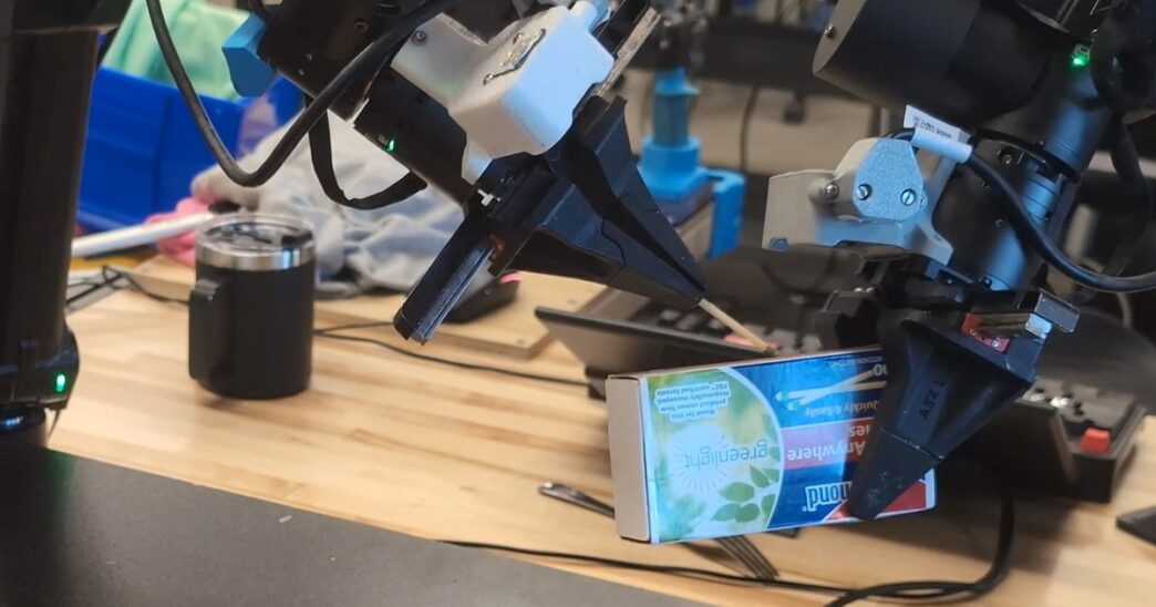 Two robot arms with black parallel-jaw grippers and wrist cameras striking a match against a matchbox on a wooden lab workbench