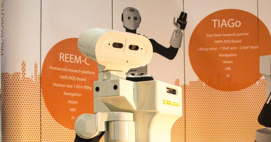 PAL Robotics TIAGo mobile manipulator robot at a RoboCup exhibit stand, with a REEM-C humanoid robot behind it
