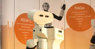 PAL Robotics TIAGo mobile manipulator robot at a RoboCup exhibit stand, with a REEM-C humanoid robot behind it