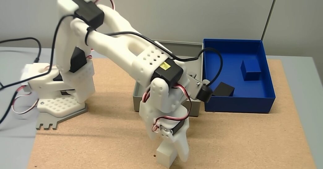 White SO-101 robot arm gripping a white block on a cork tabletop, next to a grey container and a blue container holding dark blocks