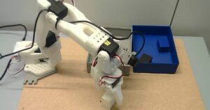 White SO-101 robot arm gripping a white block on a cork tabletop, next to a grey container and a blue container holding dark blocks