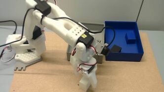 White SO-101 robot arm gripping a white block on a cork tabletop, next to a grey container and a blue container holding dark blocks